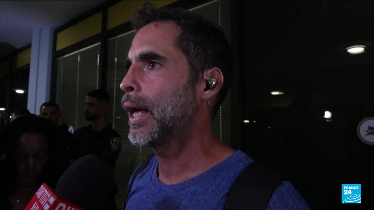 Man wearing an earpiece speaks to reporters as microphones are held near him.