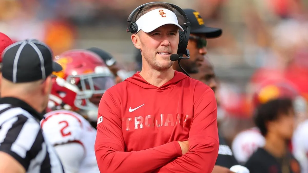 A USC coach in a red shirt and white visor stands with arms crossed during a football game.