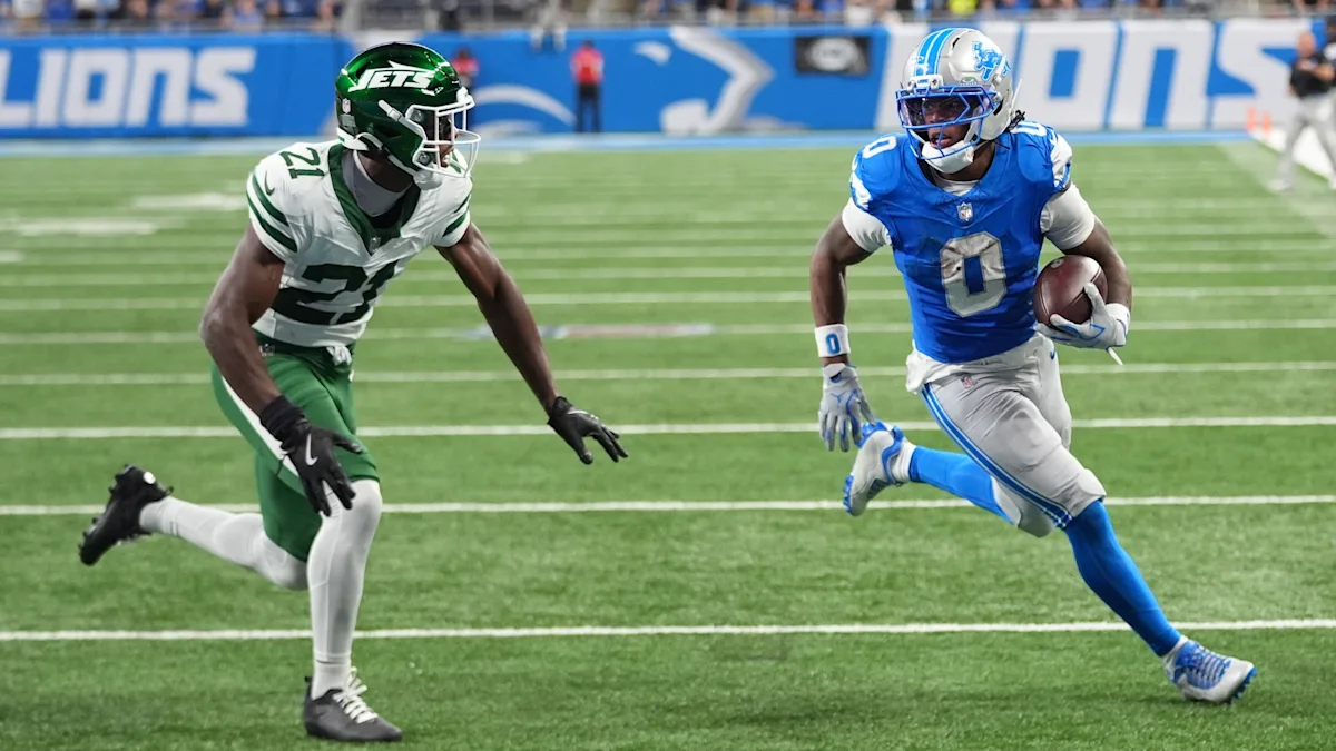 Detroit player in blue runs with the ball as a Jets defender in white and green pursues.