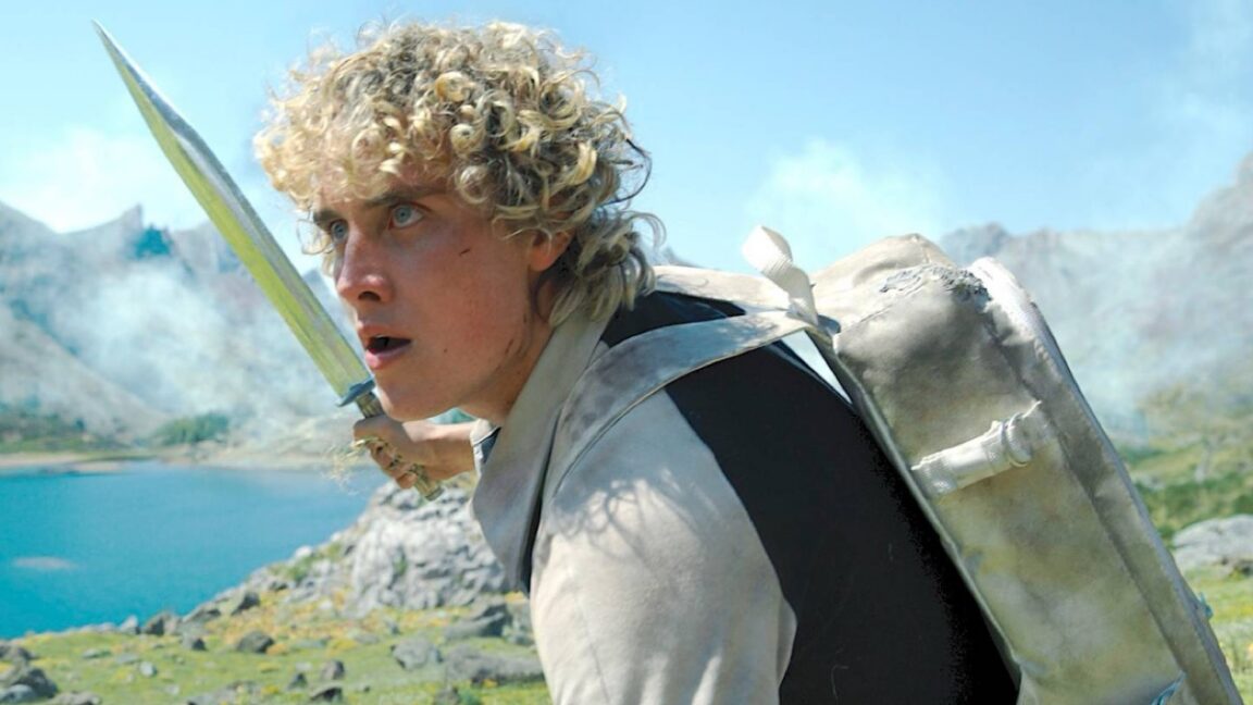 Curly-haired warrior carrying a sword and shield beside a turquoise mountain lake