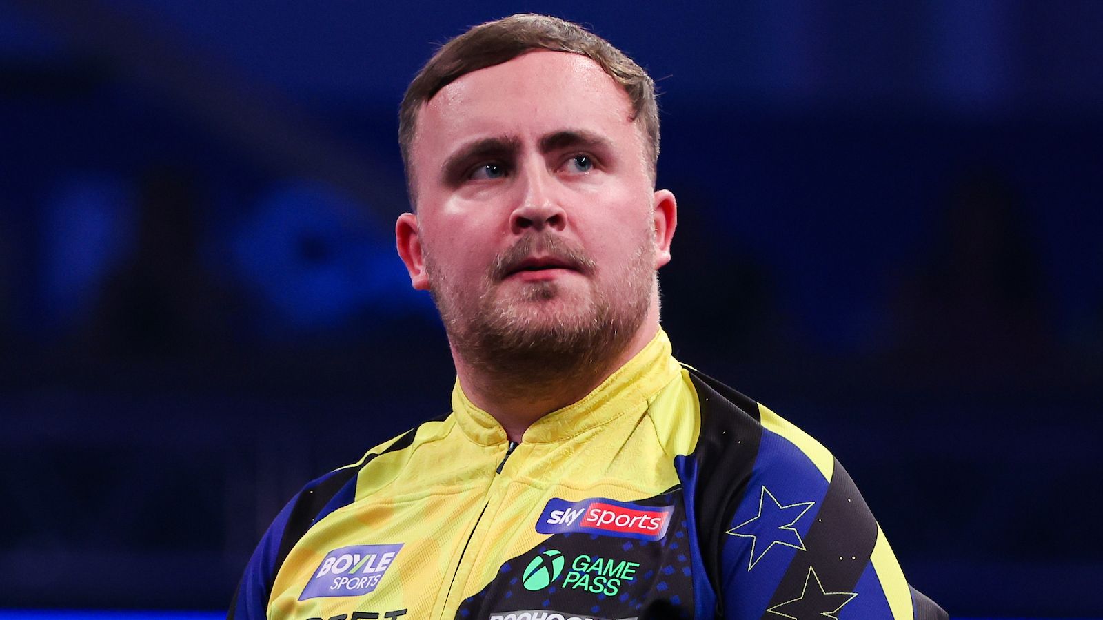 A darts player in a yellow and blue shirt looks upward against a dark arena background.