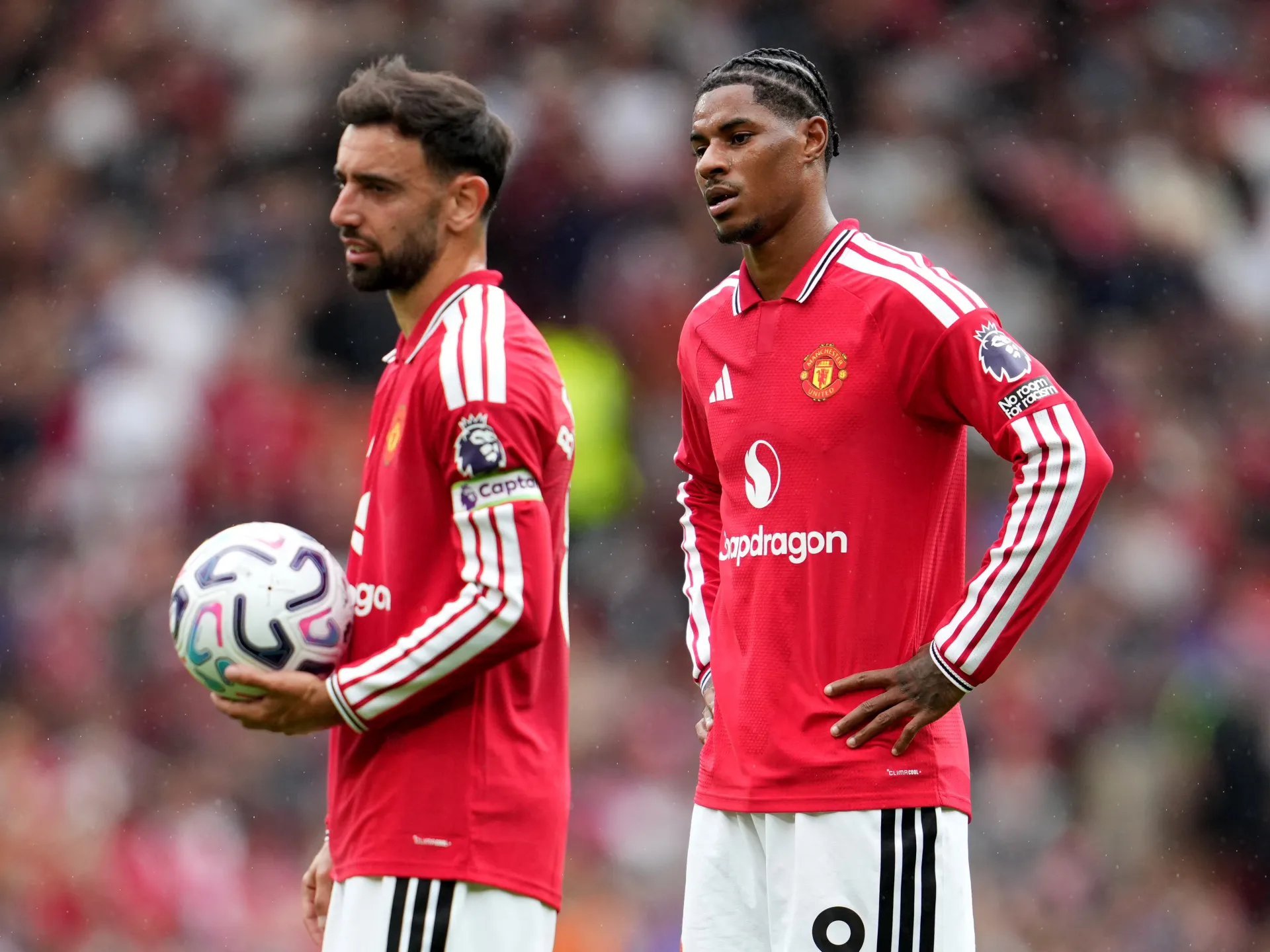 Two Manchester United players in red kits stand on the pitch, one holding a football.