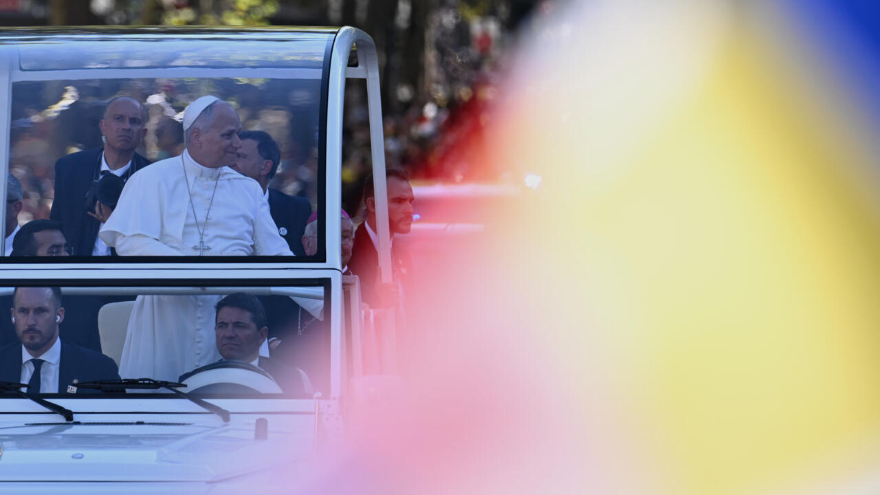 A pope in white robes rides in an open vehicle, partly obscured by blurred colored light.