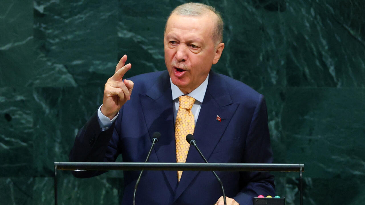 Man in a dark suit and yellow tie gestures while speaking at a lectern.
