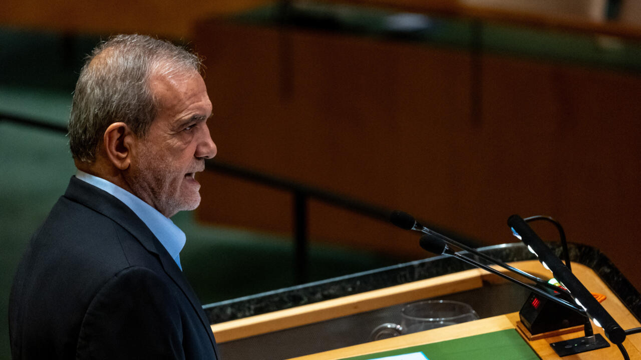 Gray-haired man in a dark suit sits beside microphones at a desk in a dim room.