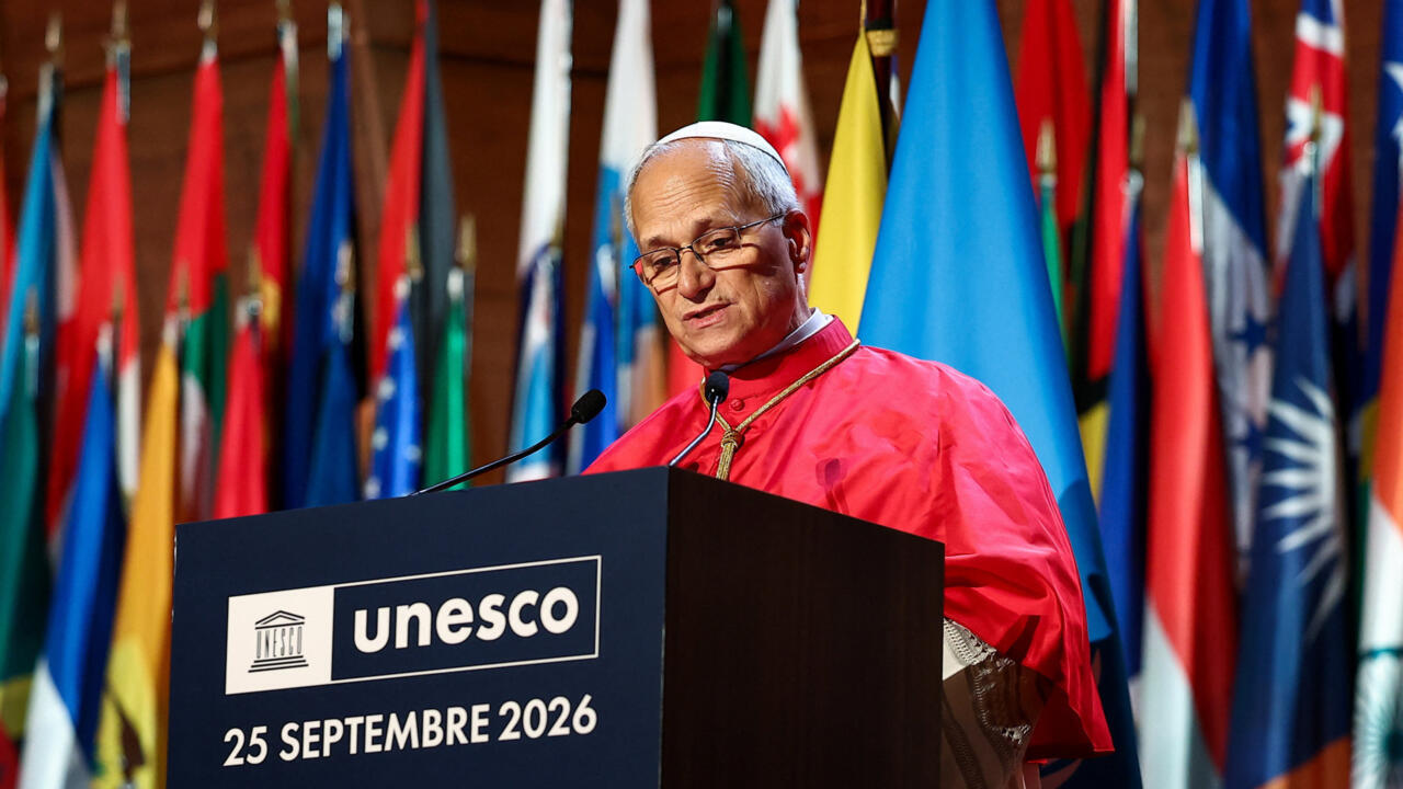 A man in red clerical clothing speaks behind a UNESCO-branded podium, with national flags behind him.