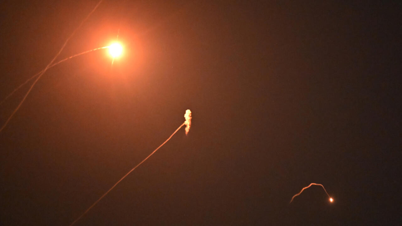 Orange lights and glowing trails cross a dark, smoky sky, with a bright flare near the upper left.