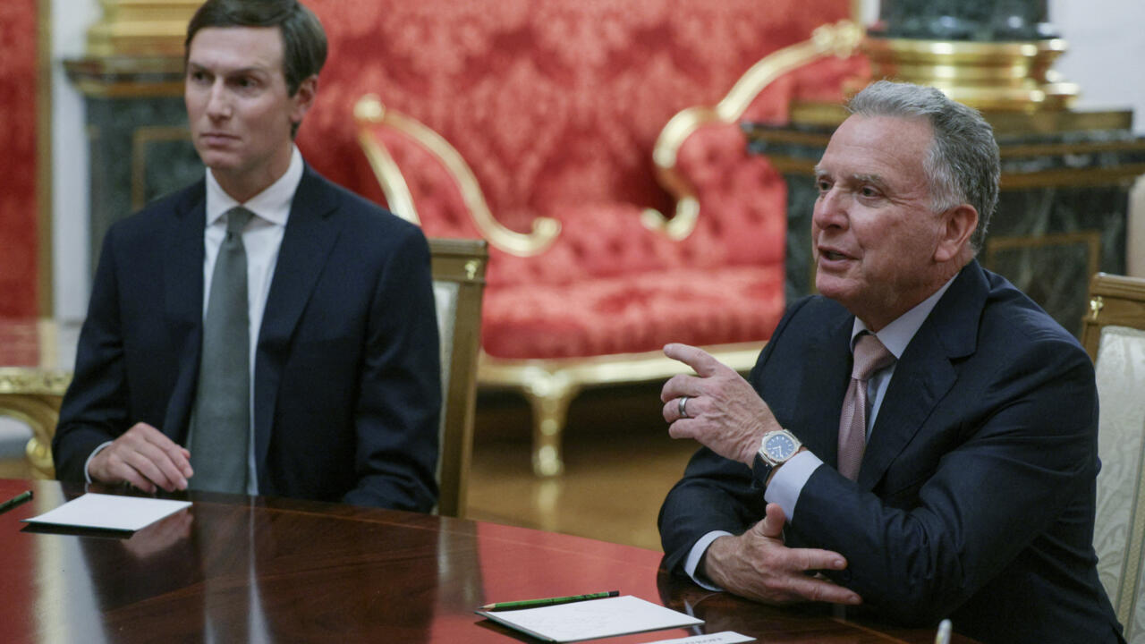 Two suited men sit at a polished table during a formal meeting, with one gesturing.