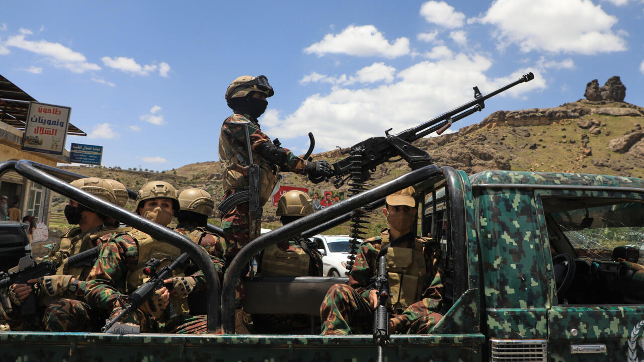 Armed men in camouflage ride in a pickup truck, one standing beside a mounted heavy machine gun.