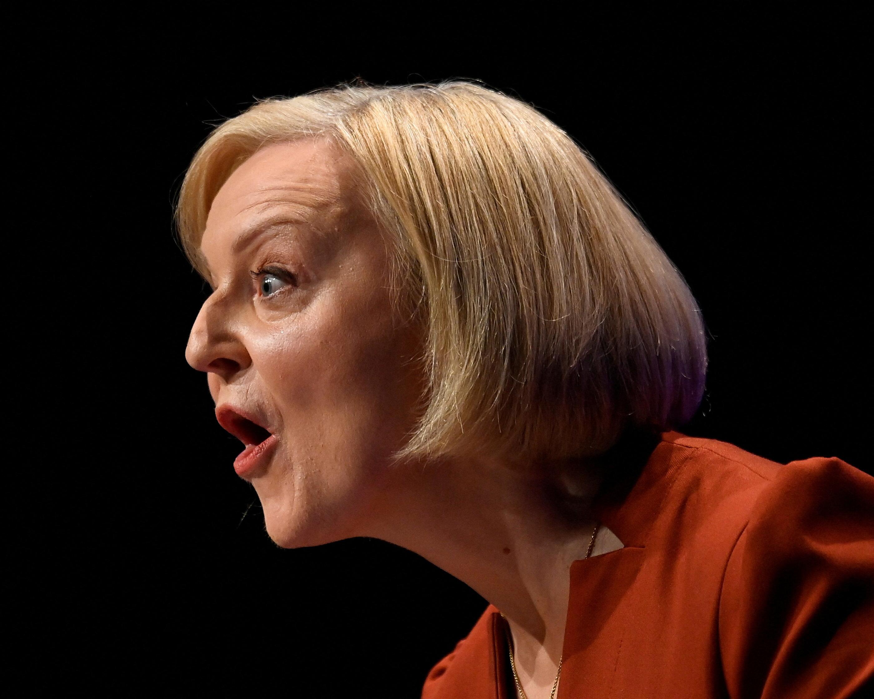 Side-profile portrait of a blonde woman speaking against a black background, wearing an orange jacket.