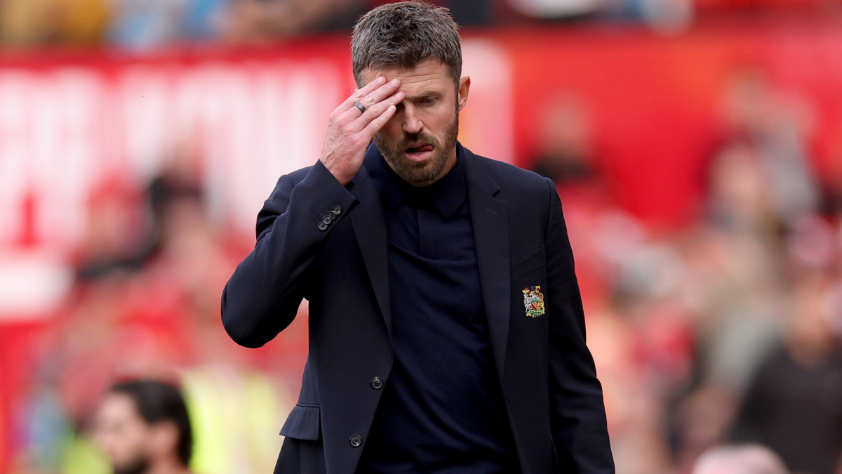 Man in dark suit touches his forehead while walking past a blurred football stadium crowd.