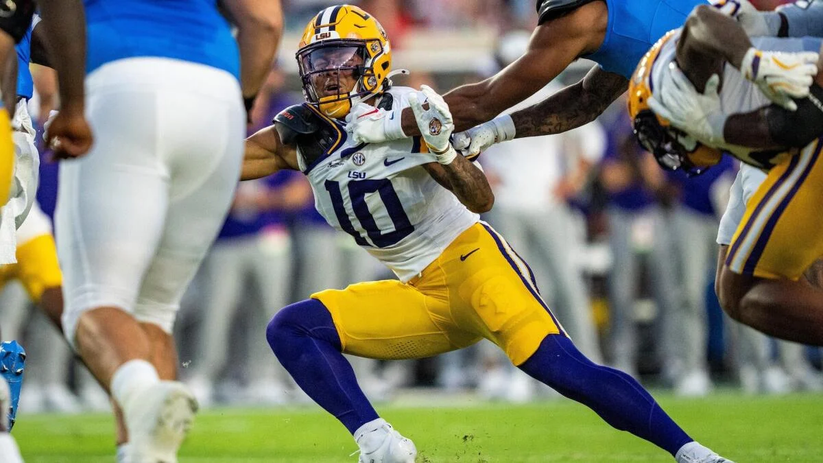 LSU football player in uniform runs with the ball amid multiple defenders.