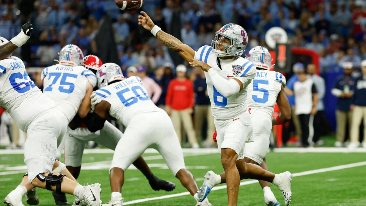 College football quarterback in white uniform throws a pass as teammates block defenders.