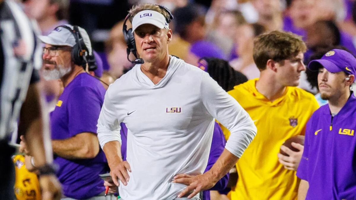 LSU coach in white visor and headset stands on sideline among players and staff