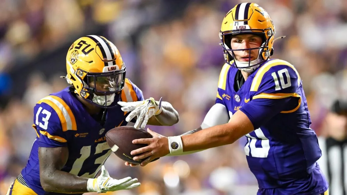 Two LSU football players in purple and gold uniforms exchange the ball during a game.
