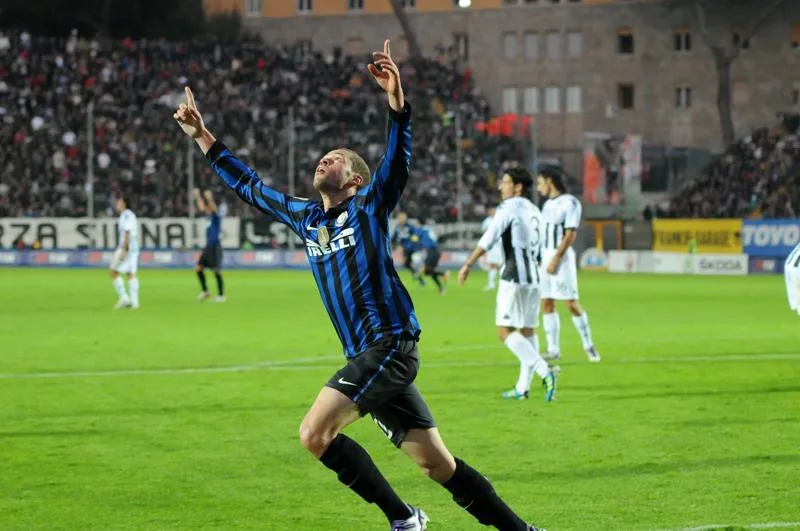 A blue-and-black striped player raises both arms as Juventus players stand on the pitch behind him.