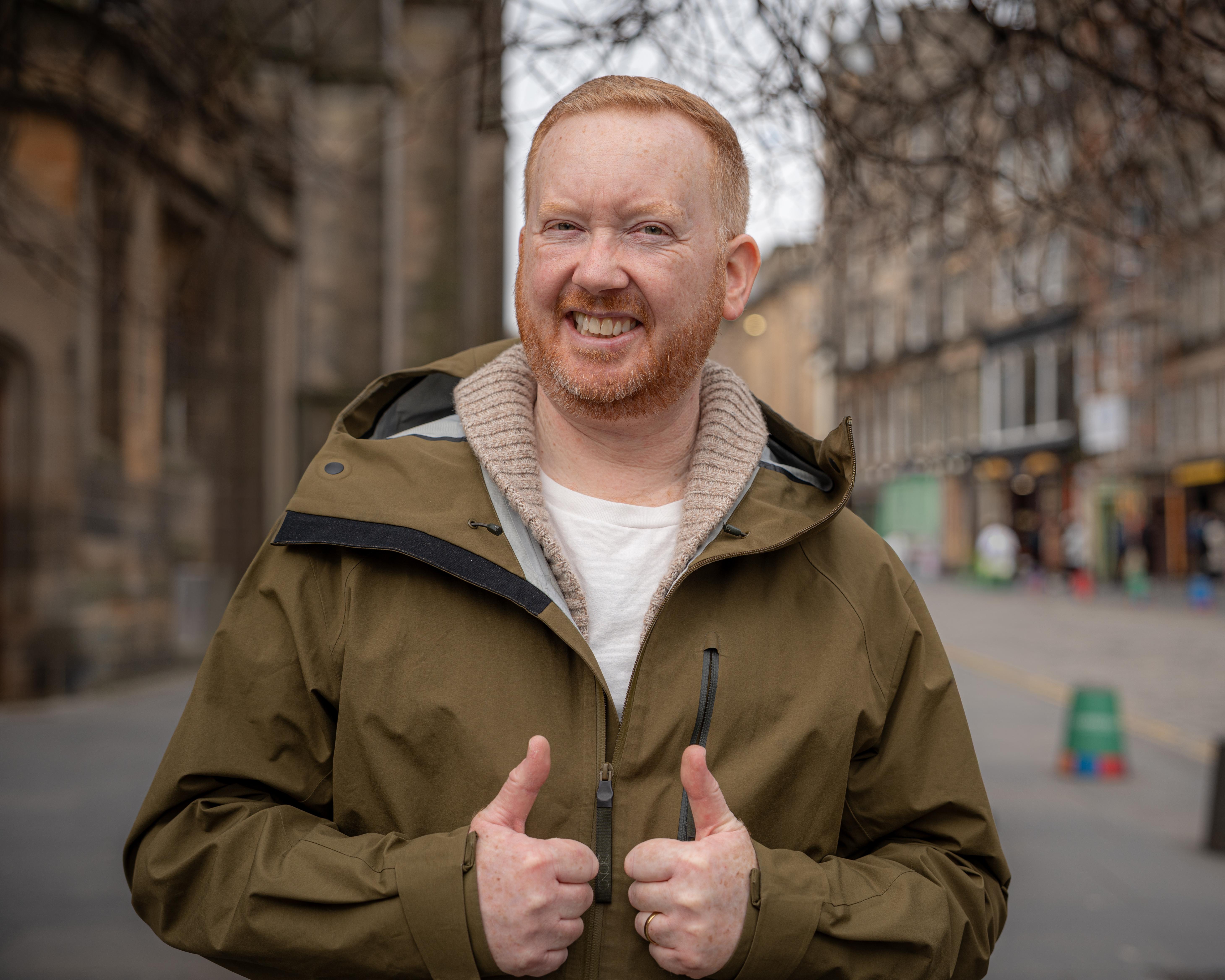 Smiling red-haired man in a brown jacket gives two thumbs up on a city street