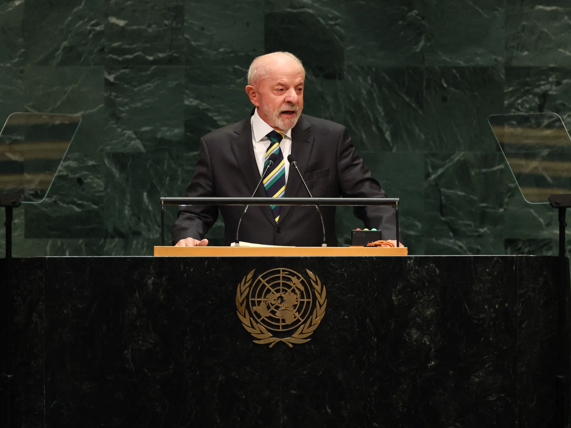Man in suit speaking at a UN podium with microphones and teleprompter screens