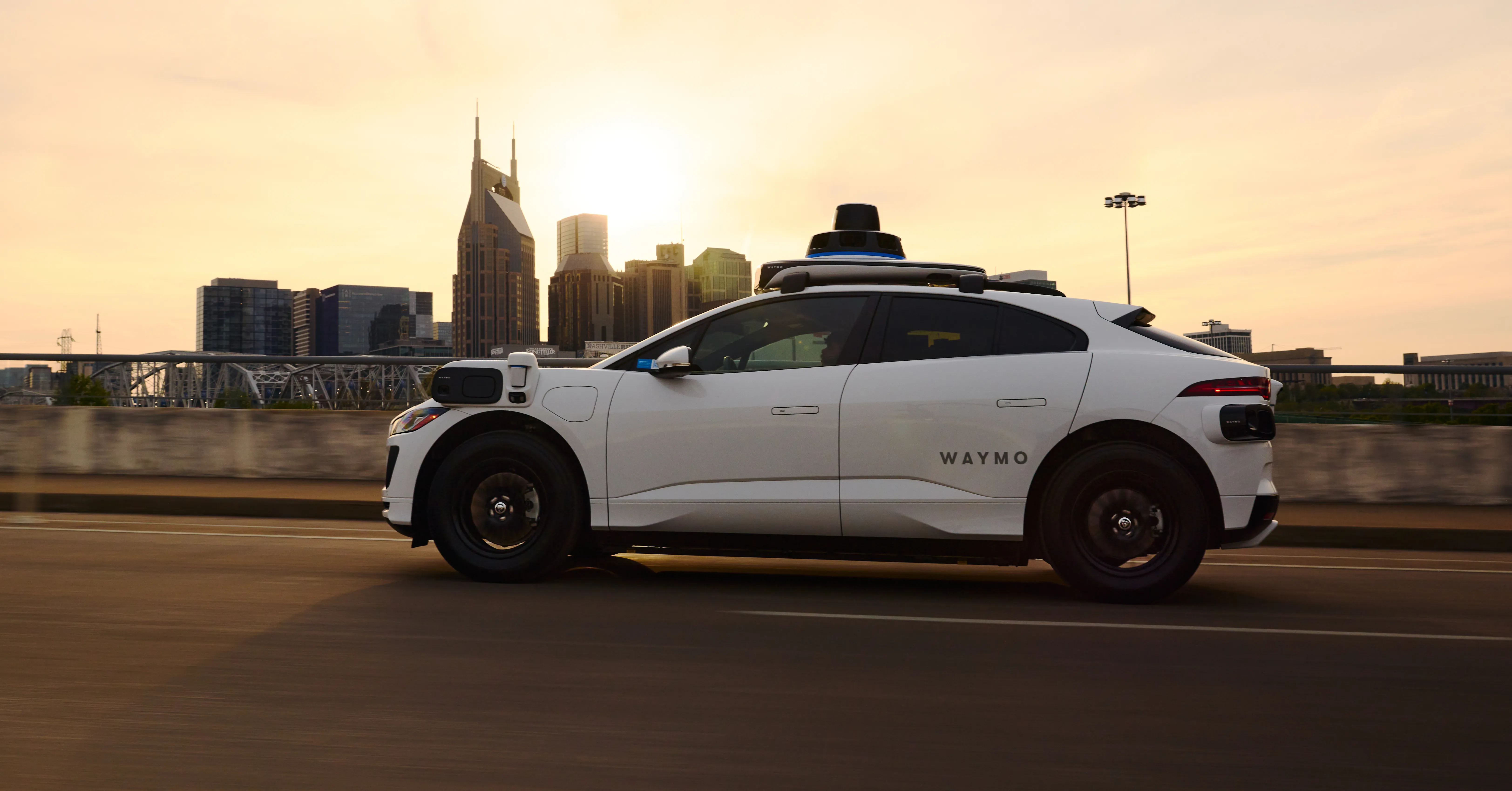 White Waymo autonomous vehicle driving past a city skyline at sunset