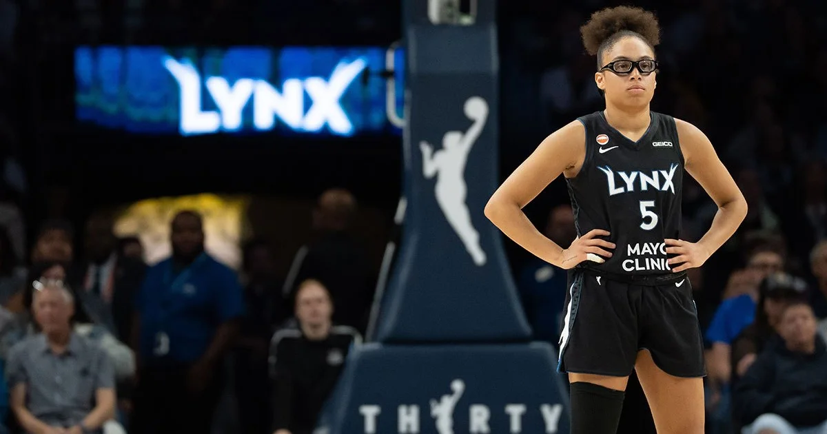 Basketball player in a black Lynx jersey stands with hands on hips near the basket.