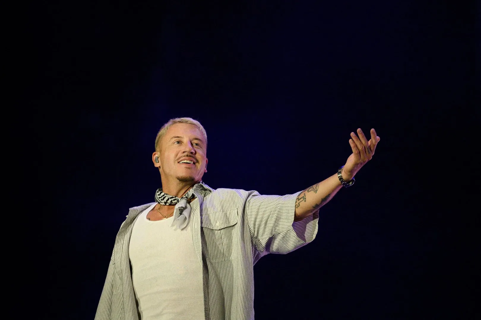 Blond male performer in striped shirt and scarf gestures with one arm raised on a dark stage