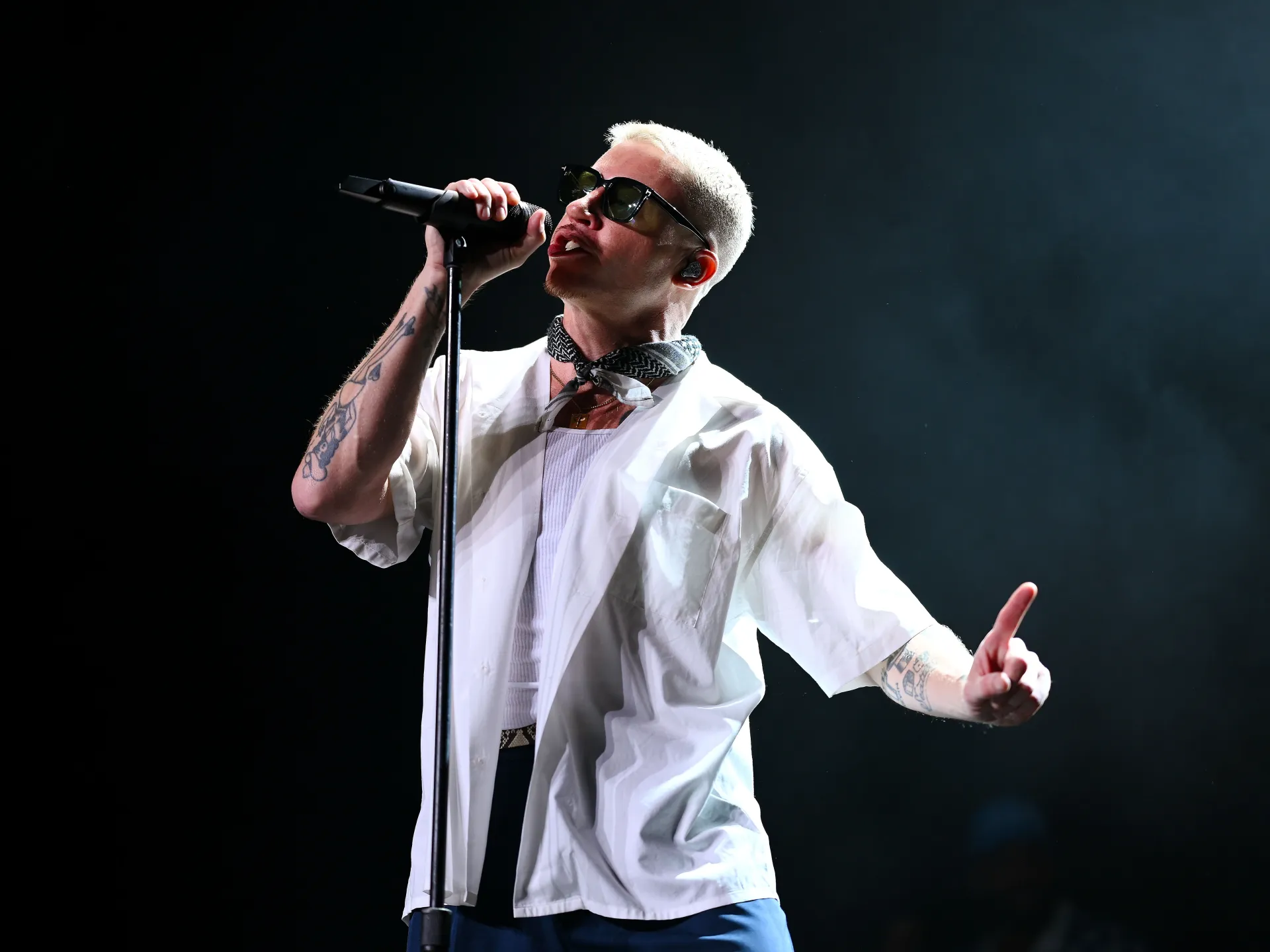 Singer with bleached hair and sunglasses performs into a microphone, extending one tattooed arm.