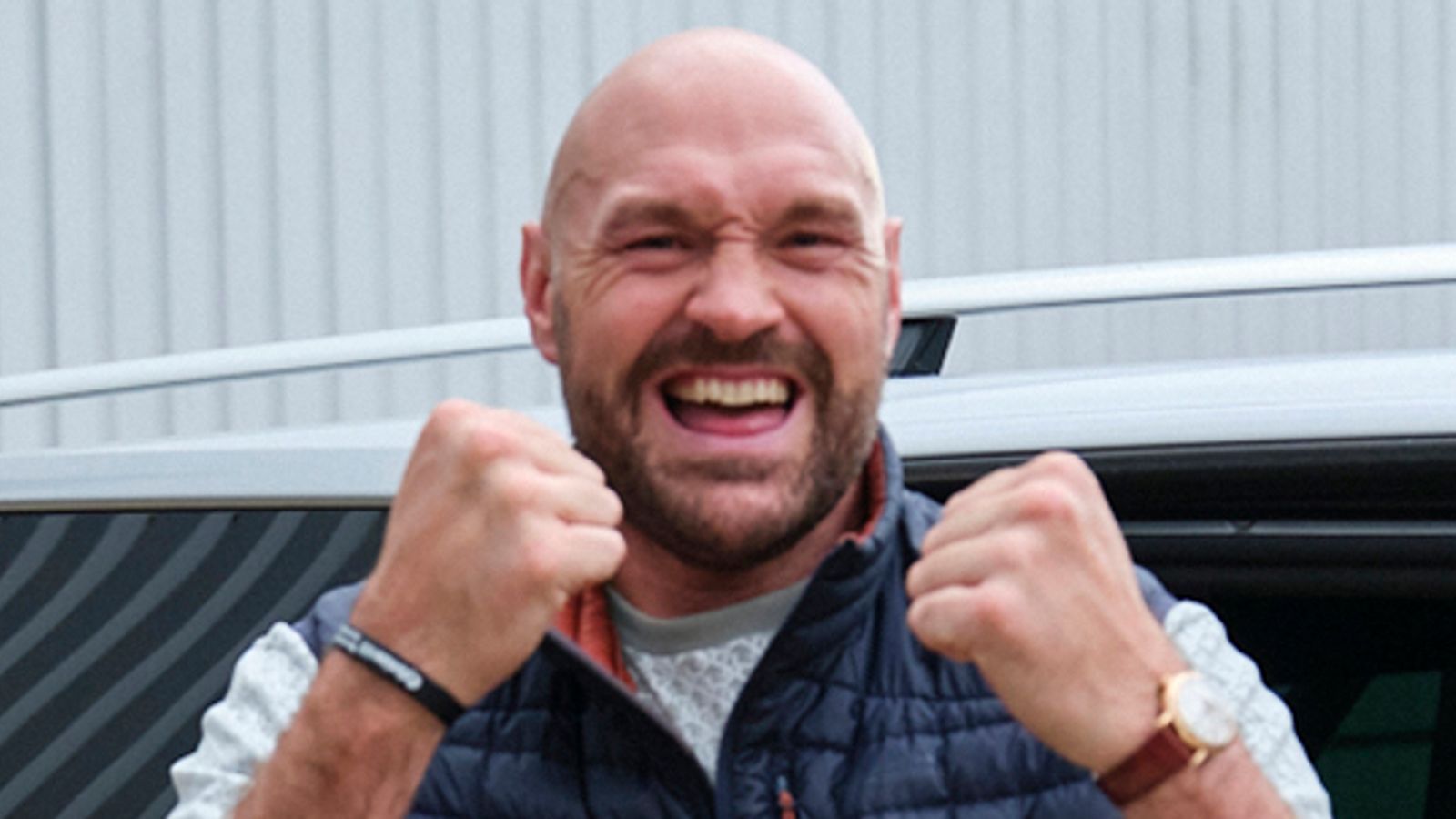 A bald, bearded man grins with both fists raised in front of a vehicle.