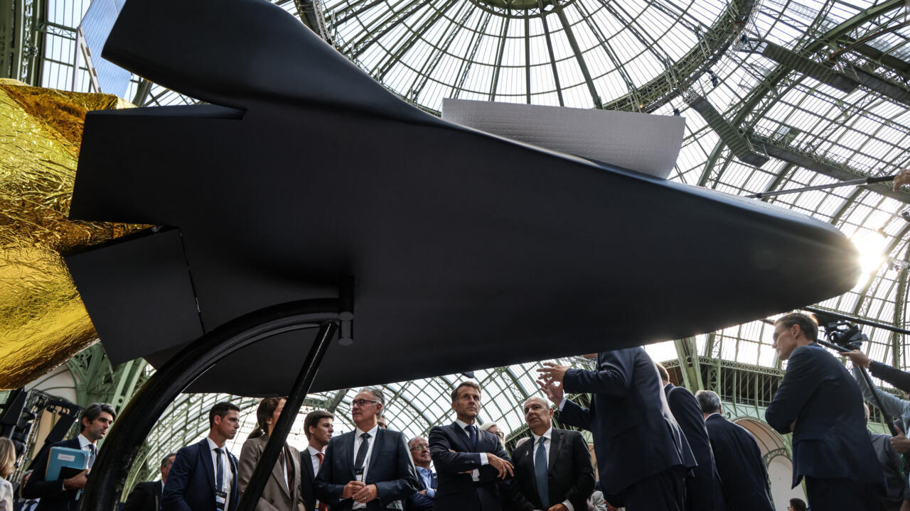 Officials and attendees gather beneath a large black aerospace exhibit inside a glass-domed hall.