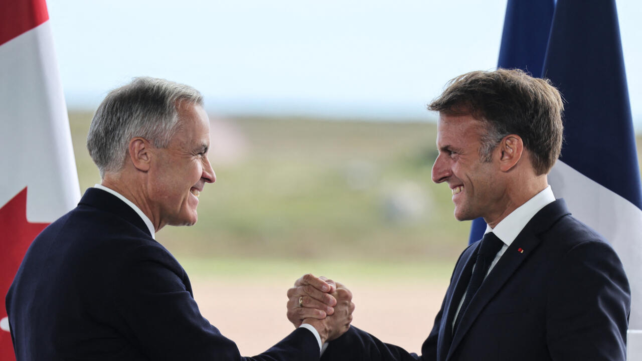Two suited men smile and clasp hands in front of Canadian and French flags.