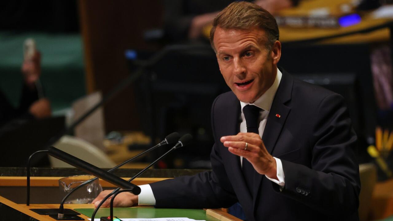 Man in dark suit gestures while speaking at a conference podium with microphones.