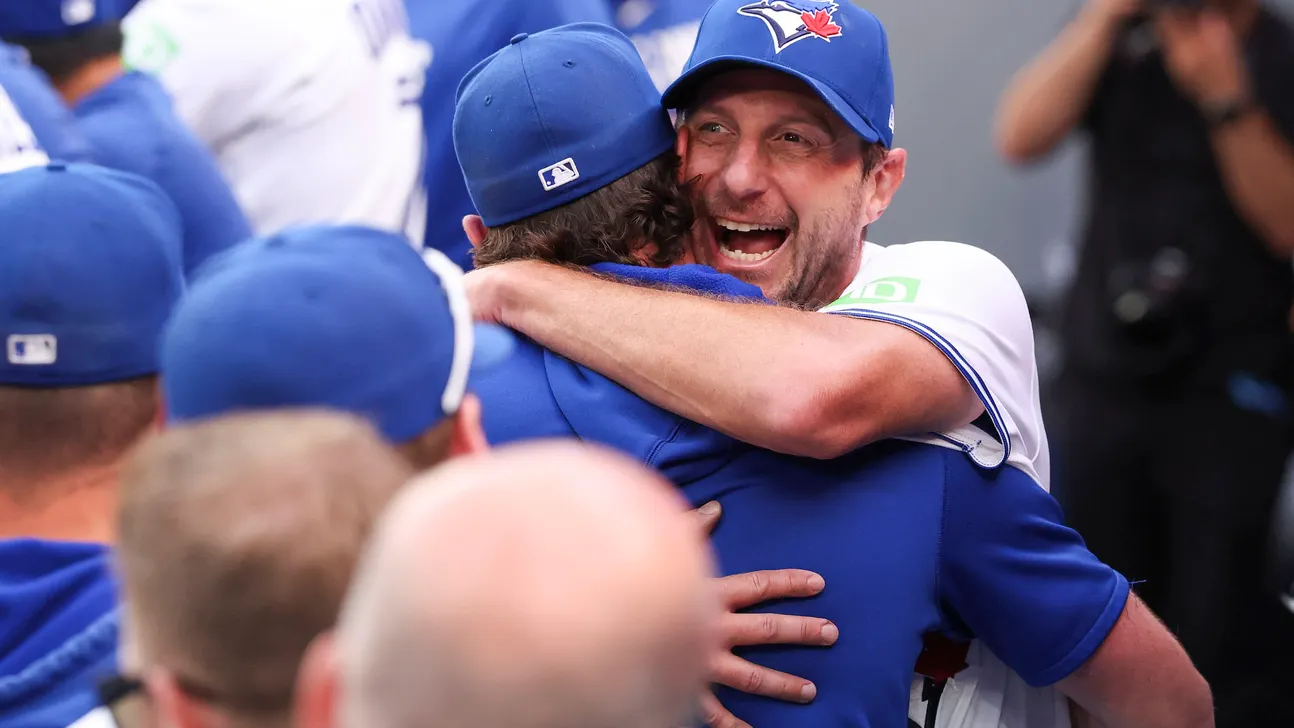 A smiling Blue Jays player embraces a teammate as others gather around them in the dugout.