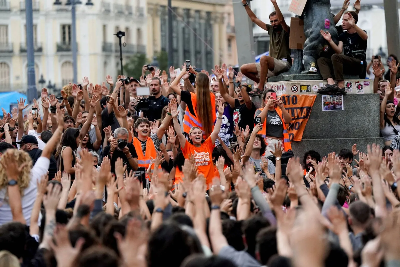 A dense crowd raises hands around a monument, with several people wearing bright orange vests.