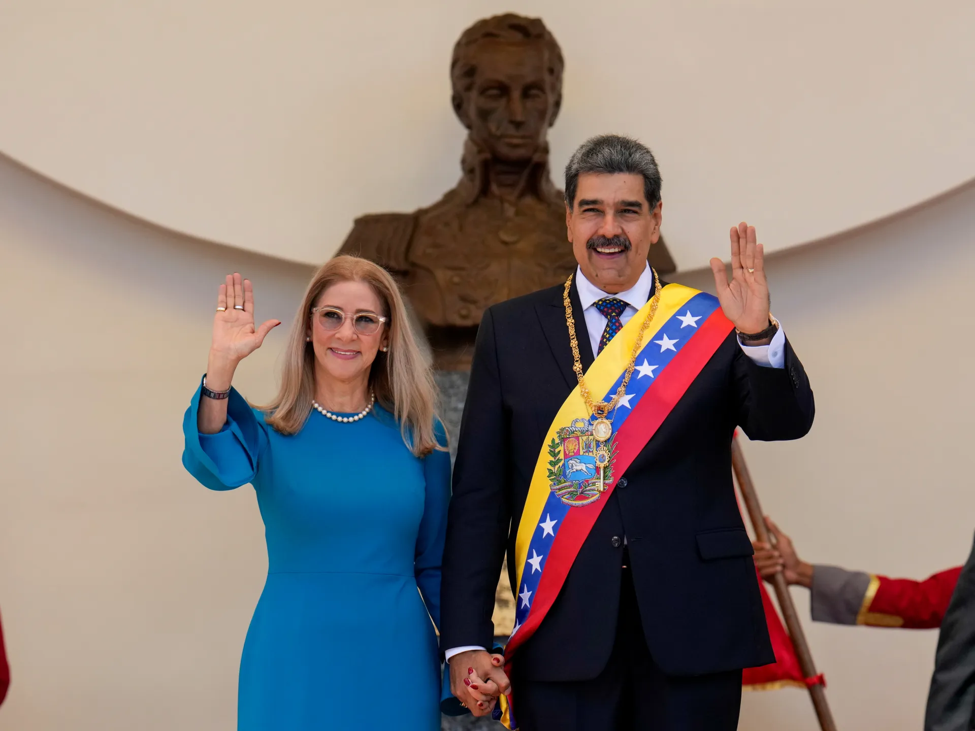 Man in ceremonial sash and woman in blue dress wave while holding hands before a large statue.