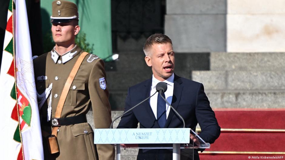 Man speaking at a podium beside a uniformed flag bearer outside a building