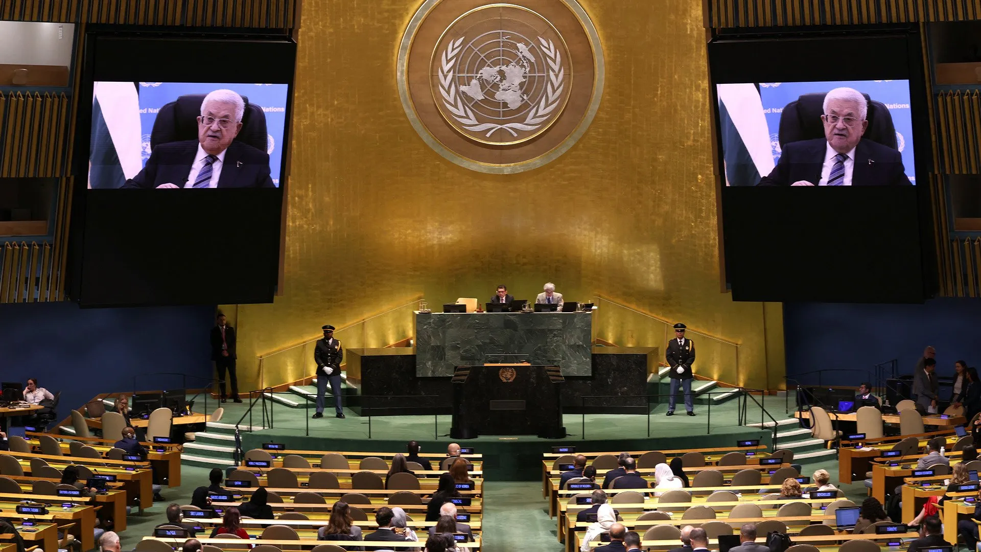 Delegates sit in a formal assembly hall as Mahmoud Abbas appears on two large screens.
