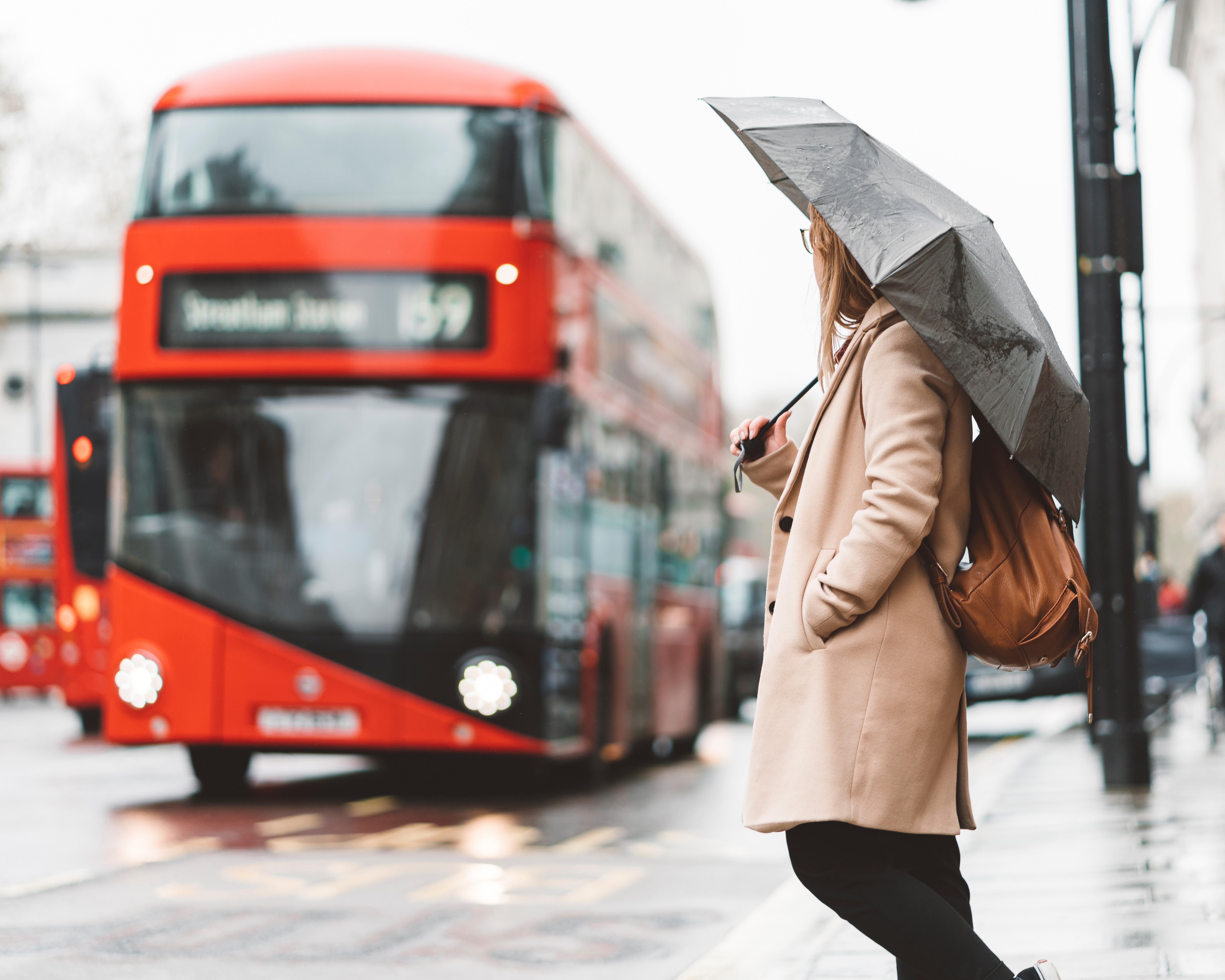 A person in a beige coat holds a black umbrella beside a red bus on a wet street.