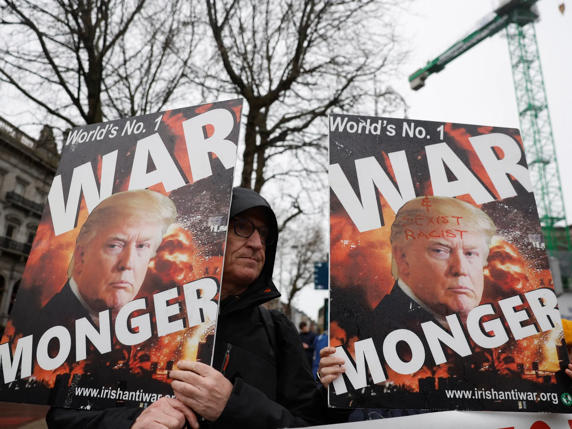 Protester holds anti-war posters featuring Donald Trump beneath leafless trees and a construction crane.