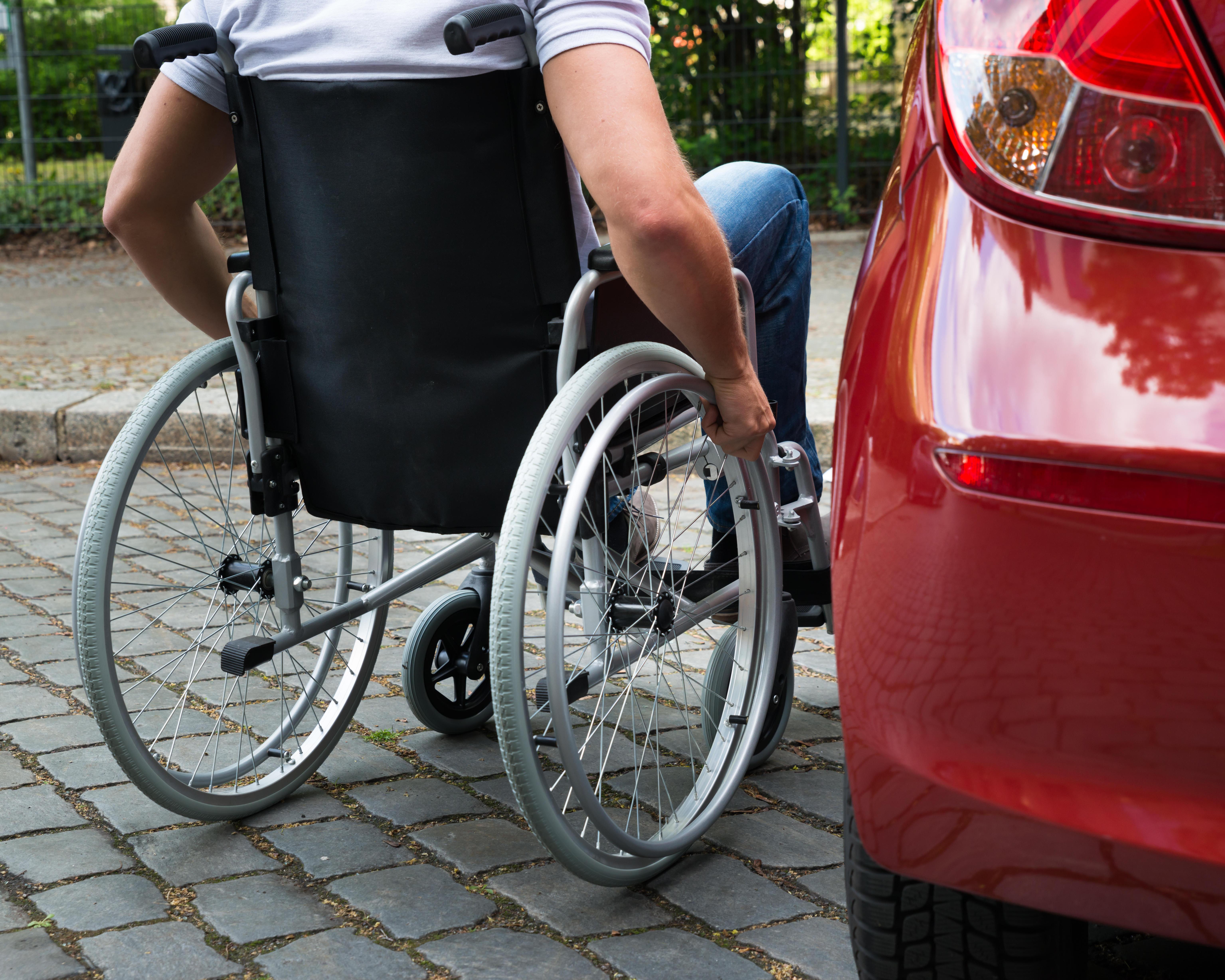 Person using a wheelchair beside a red car on a paved residential street