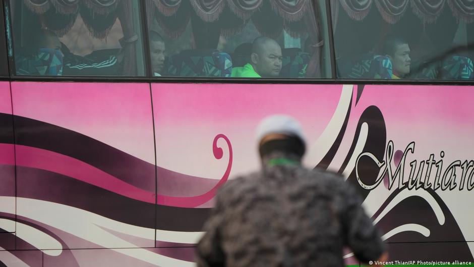 A blurred soldier stands before a pink bus with passengers visible through its curtained windows.
