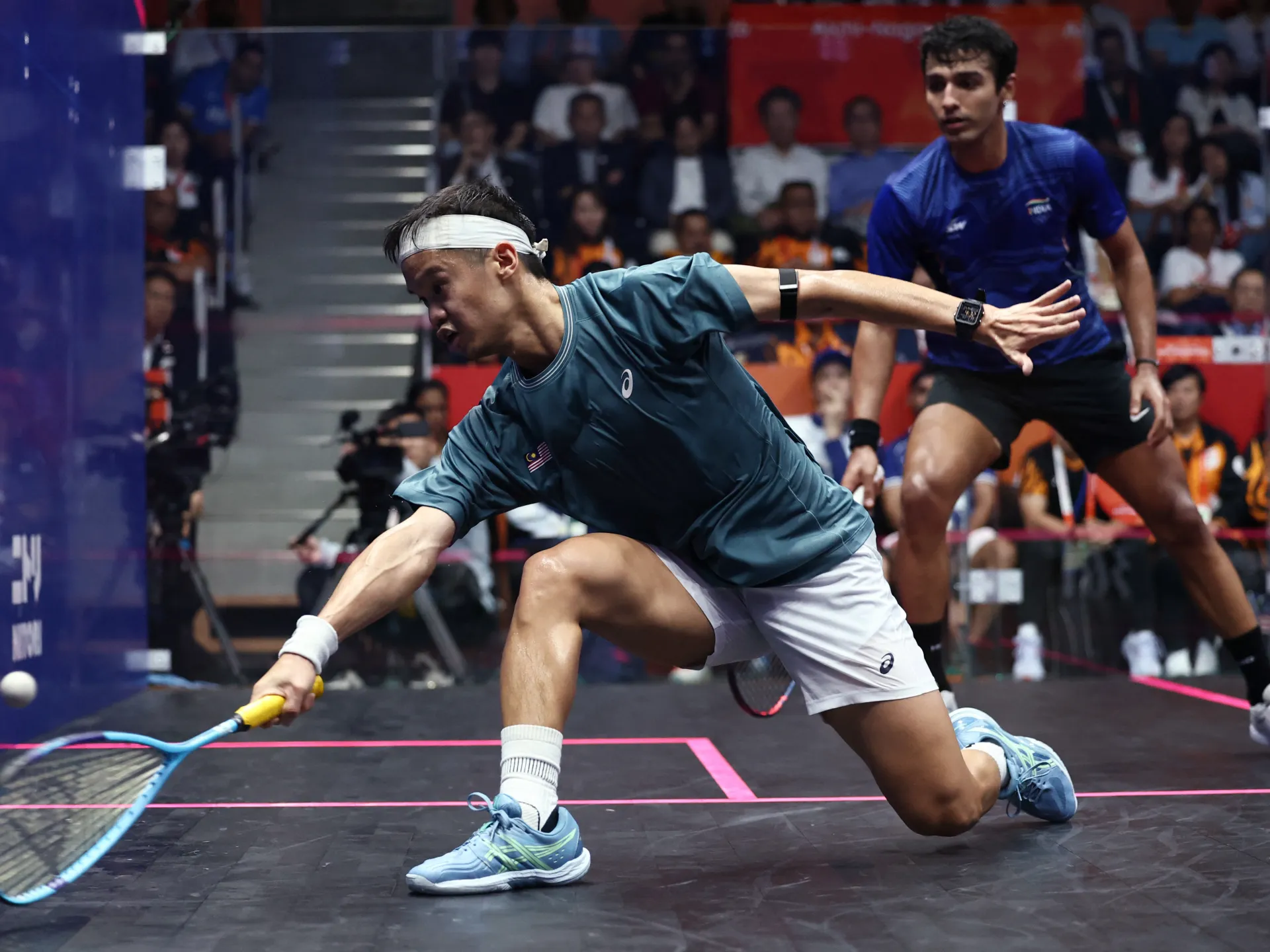 A squash player lunges low to reach the ball as his opponent watches behind him.