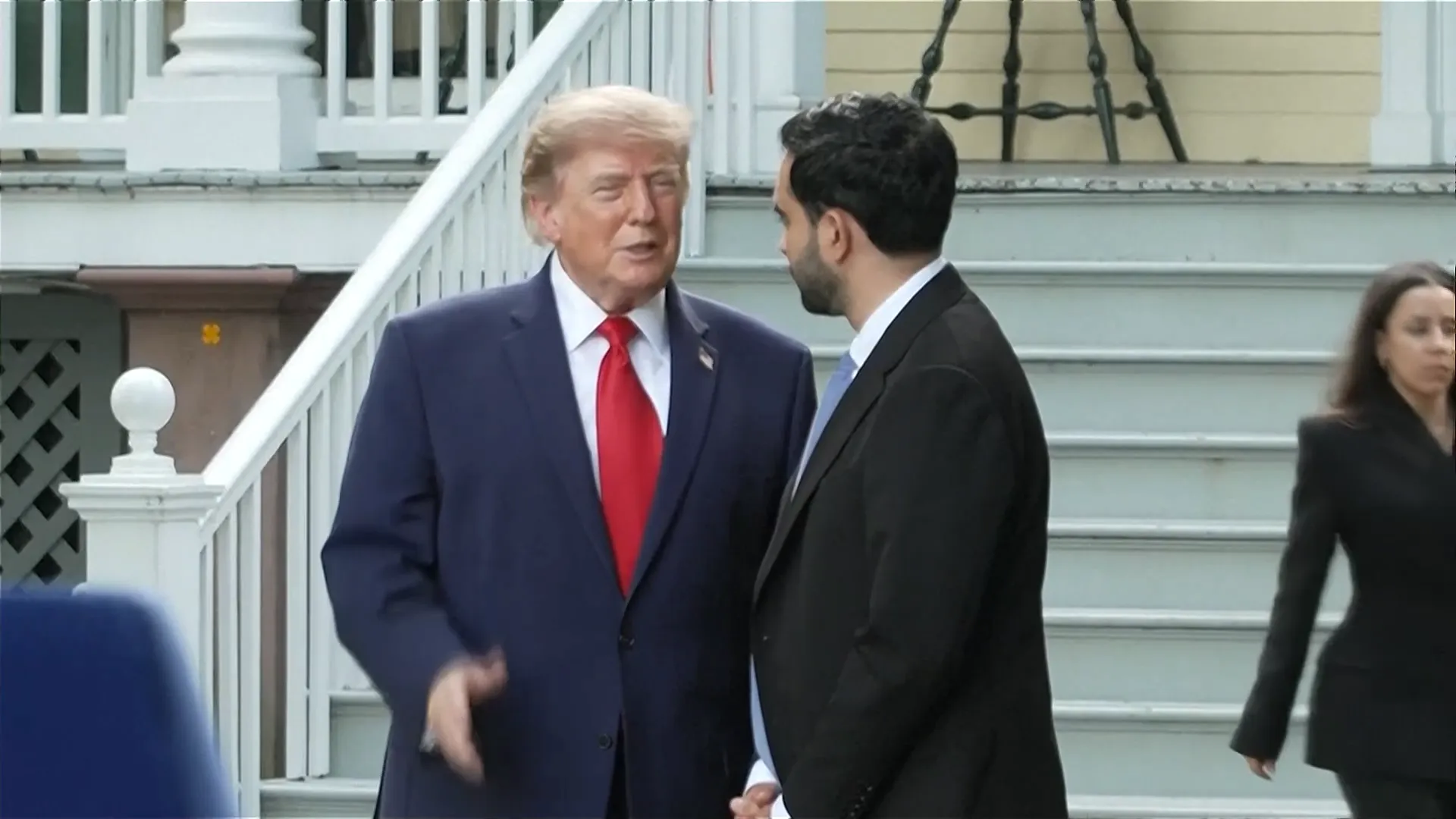 Two suited men converse outside a building as a woman walks past in the background.