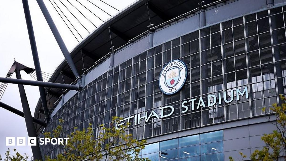 Exterior of a glass-fronted stadium with a Manchester City crest and large ETIHAD STADIUM lettering.
