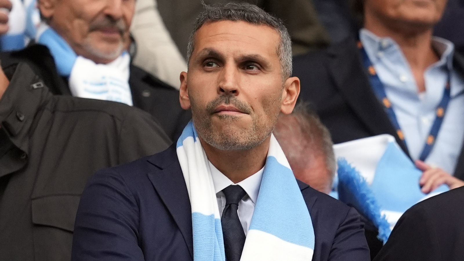 A man in a dark suit and sky blue and white scarf sits among spectators.