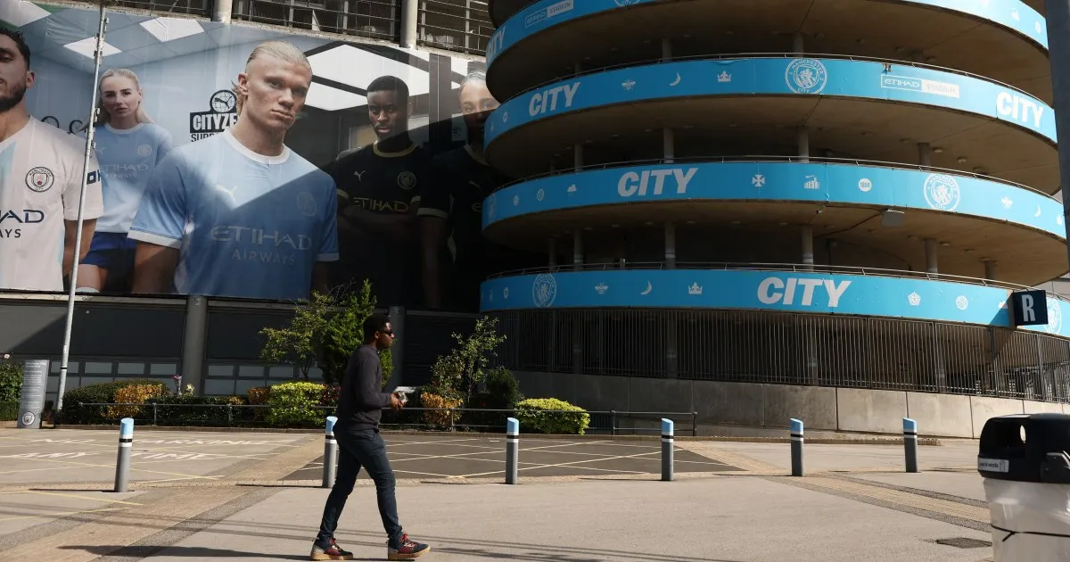 A pedestrian walks past a Manchester City building with blue club banners and footballer portraits.