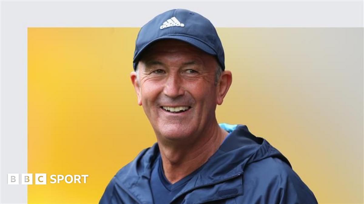 Smiling man in a navy cap and jacket against a yellow and gray background.