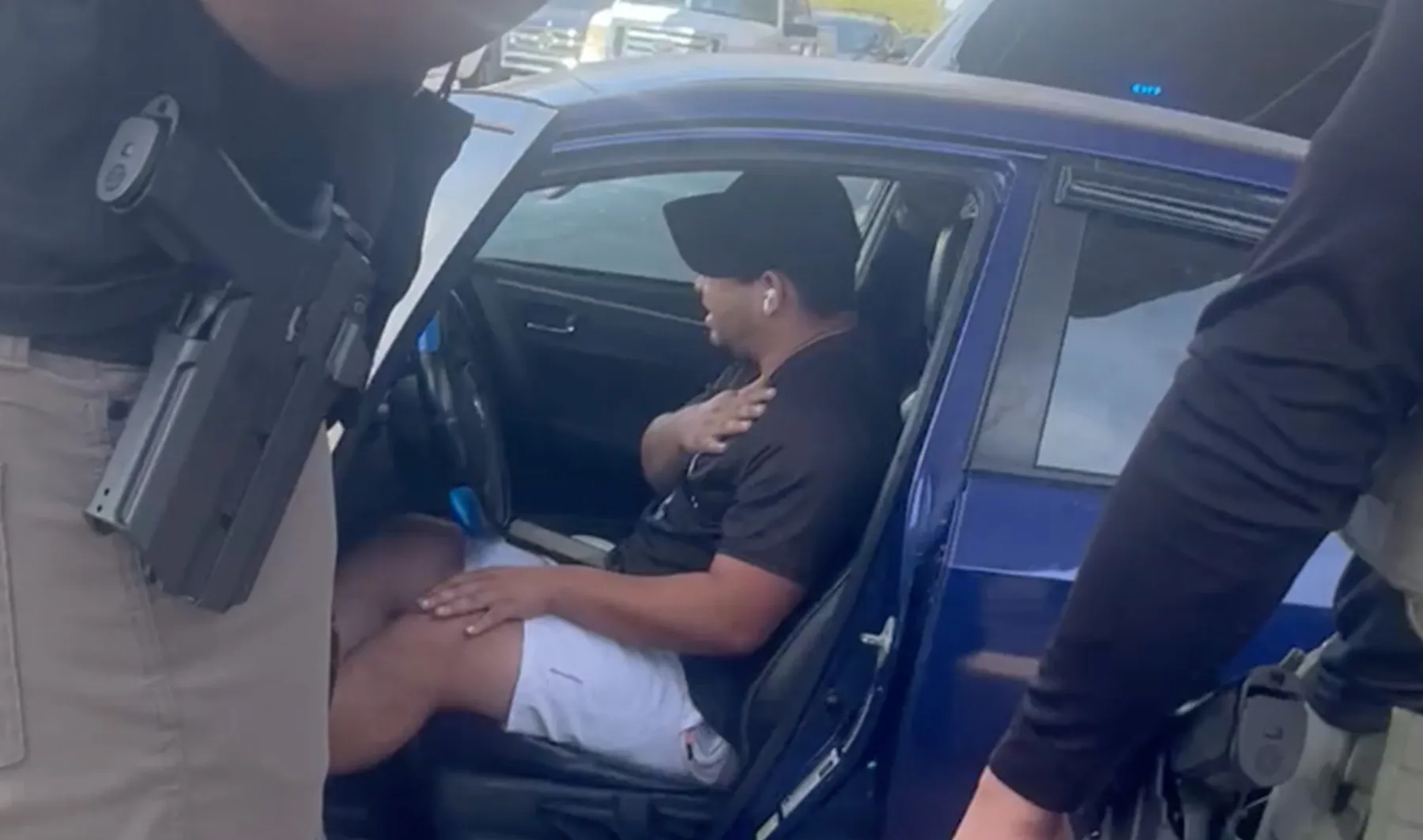 Man sits in a blue car while uniformed personnel stand nearby outside the open driver-side door.