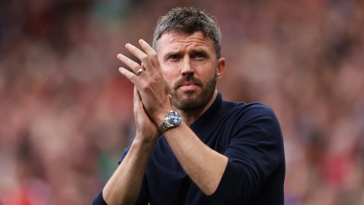 A bearded man in a dark shirt applauds with a blurred stadium crowd behind him.