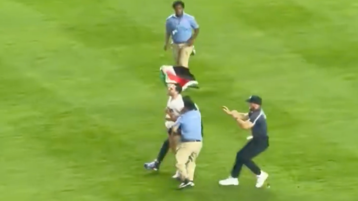 Several people run across a baseball field as one man waves a Palestinian flag.