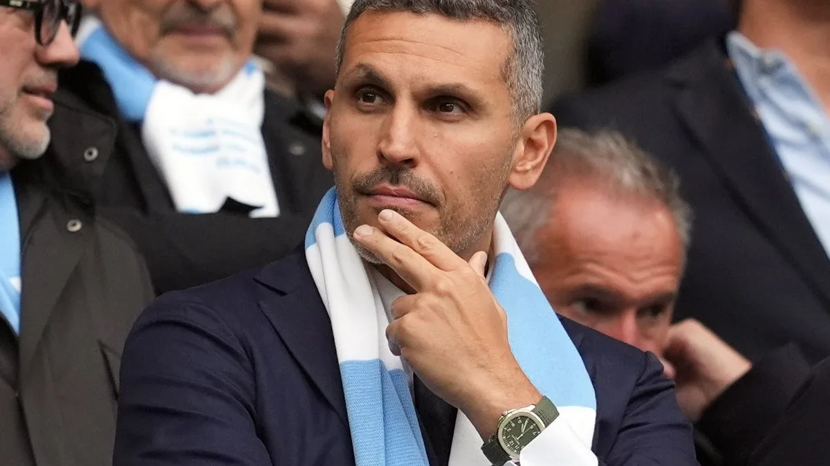 Man in a navy suit and pale blue scarf watches from the stands, fingers resting on his chin.