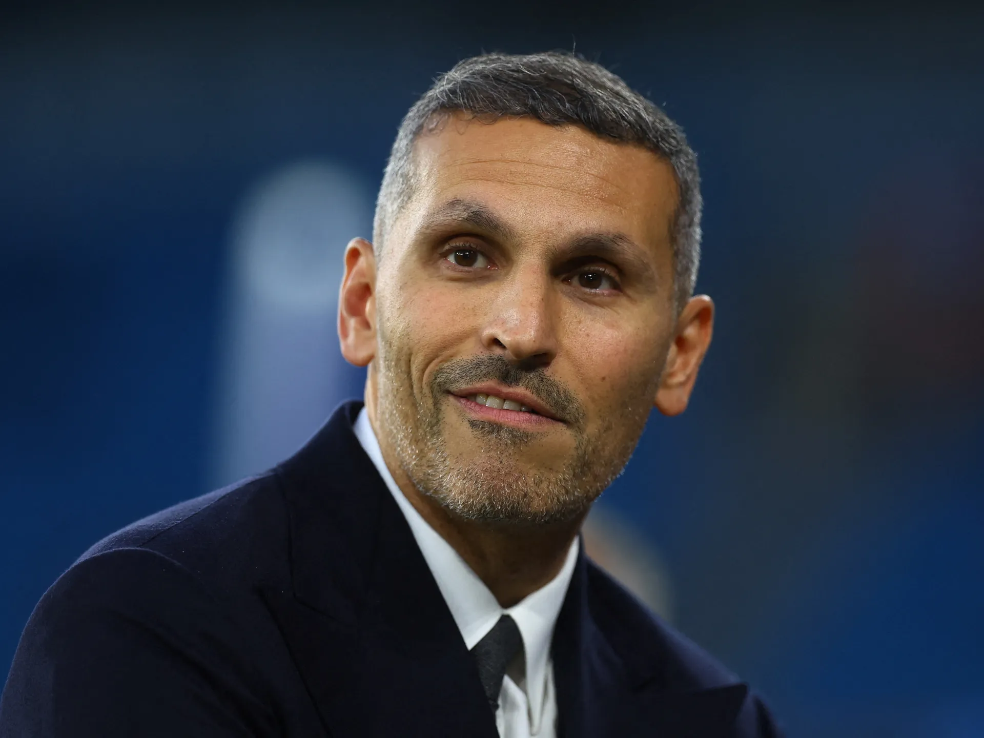 Man in a dark suit and tie against a blurred blue background.